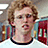 A pixelated image of a person with curly light brown hair wearing a white shirt with black collar, set against a blue background with red text.