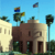 Pixelated scene shows two buildings under blue sky with flags, palm tree, and greenery.