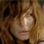Pixelated close-up of a person with light brown hair and a neutral expression, cropped to show only the upper face and hair against a dark background.