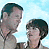 Two people stand side-by-side against a light blue sky background, with text blocks on black and gray