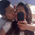 Two people pose closely together, one holding a dark rectangular object while the other leans in to kiss their cheek; background shows light-colored furniture.