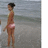 Pixelated static image of a person standing on a beach, wearing a dark cap and pink swimwear, with water and sky in the background.