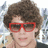 Pixelated close-up of a person with curly brown hair wearing red-framed sunglasses and a dark top against a blurred background.
