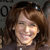 Pixelated close-up of a person with brown hair and a light-colored top, smiling with eyes partially closed against a dark background with faint text.