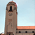 Pixelated image of a tall stone tower with a clock face, arched window, and decorative stonework against a light blue sky.