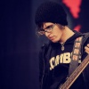 Pixelated image of a person wearing a dark beanie, glasses, and a black hoodie with yellow text, holding a guitar against a dark background with red accents.