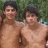 Two shirtless individuals stand side-by-side outdoors with green foliage behind them, next to a blue displaying pixelated text
