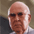 Pixelated static image of a bald elderly man with white hair on the sides, wearing dark sunglasses and a light-colored shirt against a muted brown background.