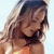 A pixelated close-up of a person with long light brown hair wearing a dark bikini top and orange garment, set against a light blue background.