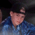 Pixelated static image of a person with light skin tone wearing a blue patterned shirt against a dark background.