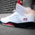 White high-top sneaker with red and black accents, featuring a visible number '23' on the side, positioned against a textured background.