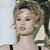 Pixelated close-up of a person with light brown hair styled in an updo, wearing a white sleeveless top against a light background.