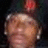 A person wearing a dark cap with a red flower, light-colored earrings, and a dark background.