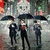 Three figures stand under umbrellas in a rainy scene; the central figure's red tie is visible, and the background includes a large moon-like shape.