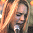A woman with long blonde hair holds a microphone, transitioning from looking down to smiling while holding the mic.