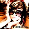 A person with brown hair and sunglasses poses with hands near their face, wearing a dark top with a light blue bracelet, against a blurred background with orange and brown tones and the text "PERFECTION" at the bottom.