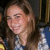 A person with long brown hair wears a blue and white patterned top, smiling in three showing slight head movement
