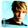 Pixelated portrait of a person with light brown hair against a light blue background, wearing a dark shirt with visible text.