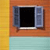A static pixelated image shows a window with open shutters against a wooden wall, with a yellow vertical section on the left and a light blue lower section.