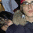 A person with light brown hair and glasses wears a dark blue top, holding hands in front of them while standing under a red cap.