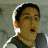 Pixelated close-up of a person with short dark hair wearing a dark jacket over a light shirt, mouth open in mid-expression against a muted background.