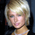 Pixelated close-up of a person with short blonde hair, light skin, and a neutral expression against a dark background.