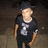 Pixelated image of a person wearing dark cap, black t-shirt with white graphic design, and dark pants against textured background.