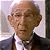 Pixelated portrait of a person with light skin tone, short light brown hair, wearing a dark suit jacket over a white shirt and dark tie, set against a light background with a dark vertical element on the right.