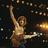 Pixelated image of a person playing guitar on stage with raised arm, surrounded by blurred lights and orange background text.