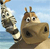 Pixelated cartoon giraffe with brown and white patches, large eyes, and small horns, standing on a sandy beach with blue water and sky background.