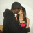 Two figures in close embrace, one wearing a dark hat and dark clothing, the other in a red top, against a light background.