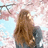 A woman with long brown hair stands under pink cherry blossoms, viewed from behind against a light blue sky.
