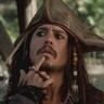 Static close-up of a man with long dark hair wearing a brown tricorn hat, holding his index finger to his lips in a shushing gesture against an outdoor background.