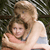 Two people embrace in a close-up pose with one person holding a small dog, set against a blurred outdoor background.