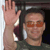 A pixelated image shows a person with short dark hair wearing orange-tinted glasses, a dark green shirt, and white earbuds, raising a light-skinned hand in a waving gesture against a dark background.