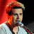 A person with light brown hair wearing a white shirt stands before a microphone against a red and black background.