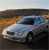 Pixelated image of a light blue sedan with dark windows and black tires, positioned on a dark road against a blurred background of orange and brown tones.