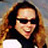 Pixelated static image of a person with long brown hair wearing dark sunglasses and a white top against a blurred background.