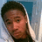 Pixelated close-up of a person with short dark hair, light skin tone, and visible facial features against a blue-gray background.