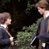 A six- shows two young men in dark blue robes with brown straps, standing outdoors among green foliage, with handwritten text overlays in yellow and blue