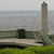 Pixelated static image showing a tall stone tower beside a body of water, with a dark rectangular structure on a concrete platform and green grass in the foreground.