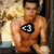 A shirtless muscular man with short dark hair sits against a textured background, wearing a black and white triangular symbol on his chest.