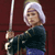 A pixelated figure with short white hair holds a long sword vertically, wearing a dark blue top and purple pants against a maroon background.