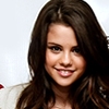 A close-up portrait of a person with long brown hair, wearing a light-colored top, against a plain light background.