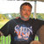 Pixelated image of a person wearing a dark shirt with a colorful graphic, standing outdoors with blurred green and yellow background.