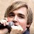 Pixelated close-up of a person with light brown hair holding a microphone near their mouth against a neutral background.