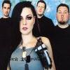 A static image shows a woman with long dark hair and heavy eye makeup in the foreground, flanked by three men in black shirts against a light blue background.