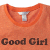A static image of an orange shirt with a white collar and silver button, featuring the text 'Good Girl' in dark brown pixelated letters at the bottom.