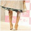 A cropped view of a person's lower legs and feet wearing a light-colored skirt with a dark patterned hem and high-heeled shoes, standing on a surface with pink and white background elements.