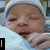 A baby wrapped in a white blanket wearing a white cap, with visible text elements and pixelated symbols on the image.